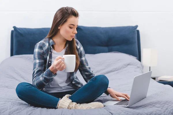 Schöne junge Frau hält Becher mit Heißgetränk und benutzt Laptop auf dem Bett — Stockfoto