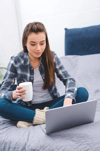 Bella ragazza sorridente tenendo tazza con bevanda calda e utilizzando il computer portatile sul letto — Foto stock