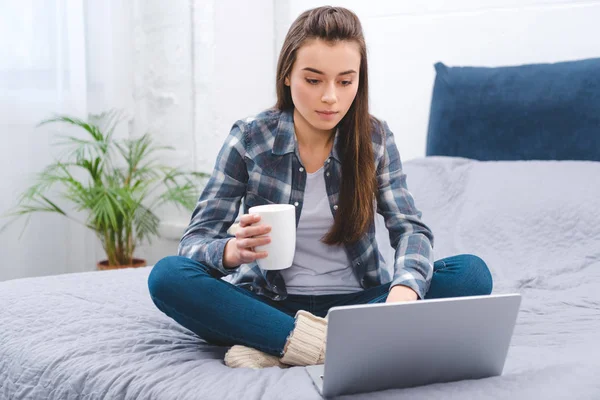 Beautiful girl holding mug with hot beverage and using laptop on bed — Stock Photo