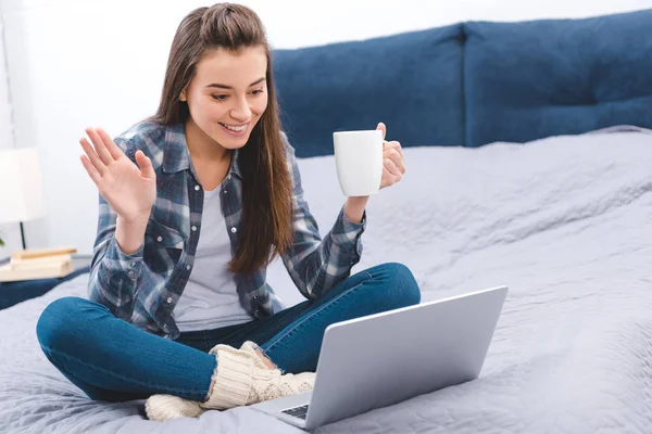 Chica feliz sosteniendo la taza y saludando la mano mientras usa el ordenador portátil en el dormitorio — Stock Photo