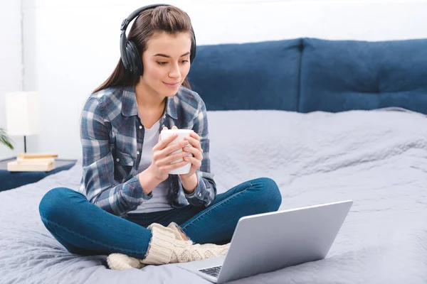 Lächelndes Mädchen mit Kopfhörer, Becher in der Hand und Laptop im Schlafzimmer — Stockfoto