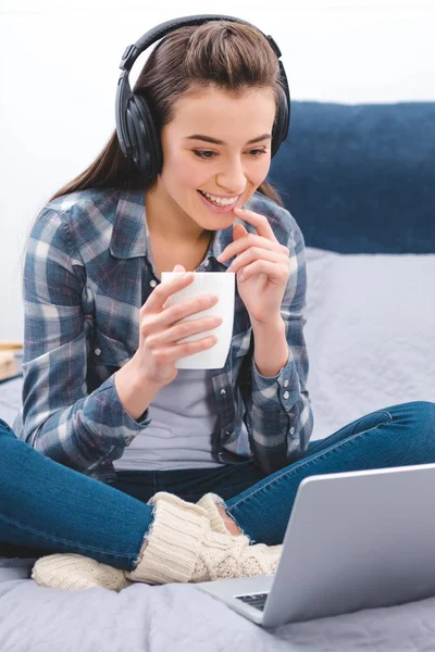 Atraente feliz jovem em fones de ouvido segurando caneca e usando laptop na cama — Fotografia de Stock