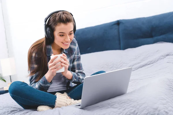 Приваблива щаслива дівчина в навушниках тримає чашку і використовує ноутбук на ліжку — стокове фото