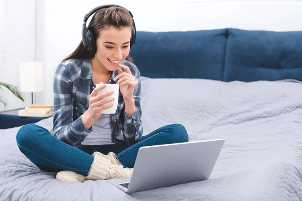 Mujer joven feliz en auriculares con taza y el uso de la computadora portátil en la cama - foto de stock