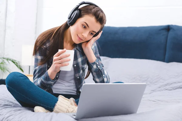 Attractive smiling girl in headphones holding cup and using laptop on bed — Stock Photo