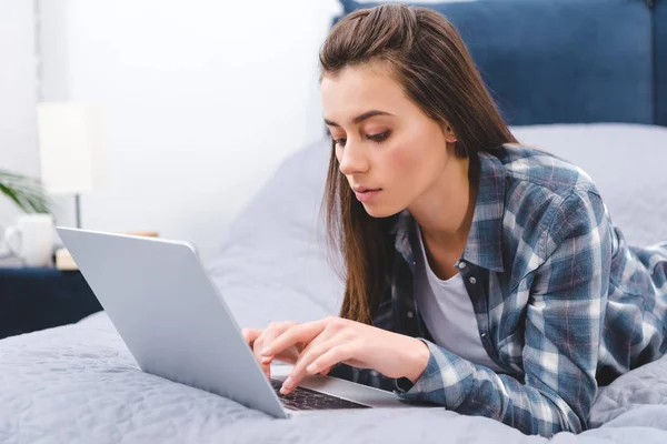 Attractive girl in checkered shirt lying on bed and using laptop — Stock Photo