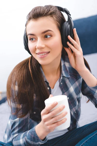 Attraente sorridente ragazza in cuffie in possesso di tazza e guardando lontano a casa — Foto stock