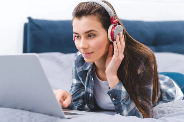 Hermosa mujer joven sonriente en los auriculares usando el ordenador portátil mientras está acostado en la cama - foto de stock