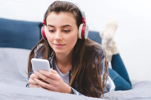 Attraktives lächelndes Mädchen mit Kopfhörern auf dem Bett liegend und Smartphone benutzend — Stockfoto