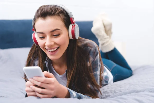 Schöne lächelnde junge Frau mit Kopfhörern, die auf dem Bett liegt und das Smartphone benutzt — Stockfoto