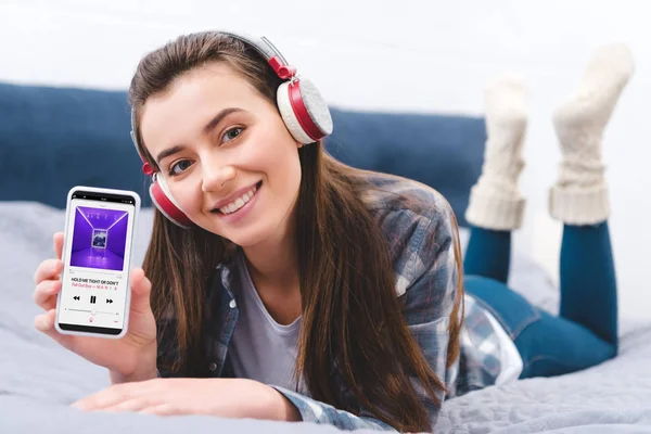 Attractive girl in headphones holding smartphone with music app and smiling at camera while lying on bed — Stock Photo