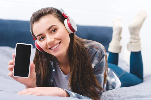 Schöne junge Frau mit Kopfhörer hält Smartphone mit leerem Bildschirm und lächelt in die Kamera, während sie auf dem Bett liegt — Stockfoto