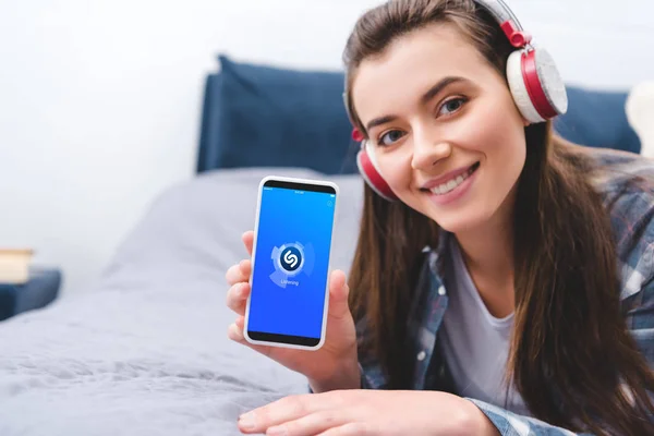 Jolie fille dans les écouteurs tenant smartphone avec application shazam et souriant à la caméra tout en étant couché sur le lit — Photo de stock