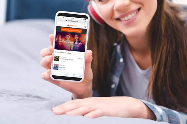 Cropped shot of smiling young woman in headphones holding smartphone with soundcloud application while lying on bed — Stock Photo