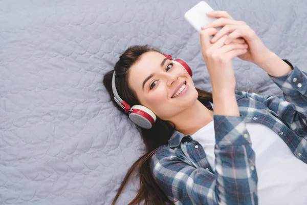Счастливая молодая женщина в наушниках, используя смартфон на кровати и улыбаясь на камеру — стоковое фото