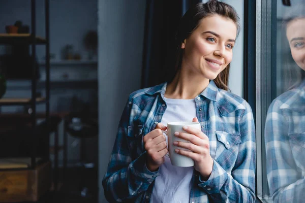 Bella felice giovane donna in possesso di tazza e guardando la finestra — Foto stock