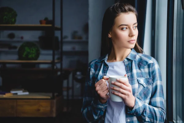 Giovane donna pensierosa che tiene la tazza e guarda la finestra — Foto stock