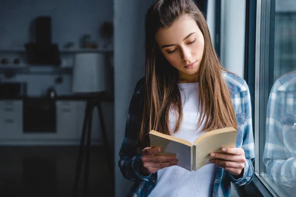 Bela jovem mulher em camisa xadrez de pé perto da janela e livro de leitura — Fotografia de Stock