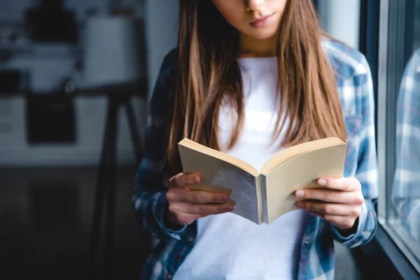 Plan recadré de jeune femme debout près de la fenêtre et le livre de lecture à la maison — Photo de stock