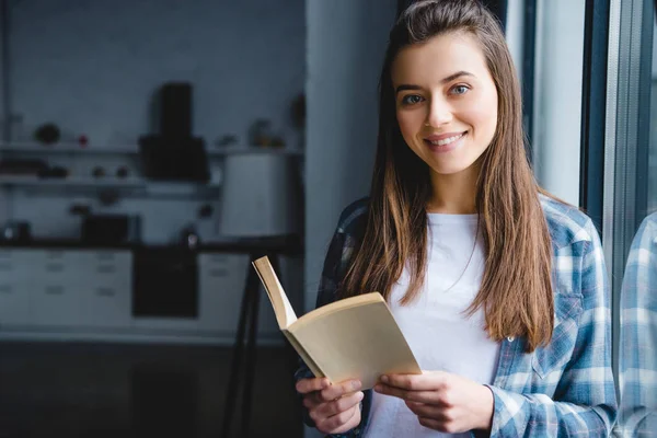 Hermosa mujer joven sosteniendo libro y sonriendo a la cámara - foto de stock