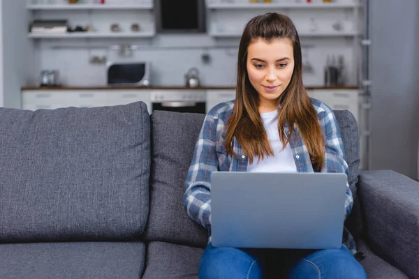 Bela sorrindo jovem mulher sentada no sofá e usando laptop — Fotografia de Stock