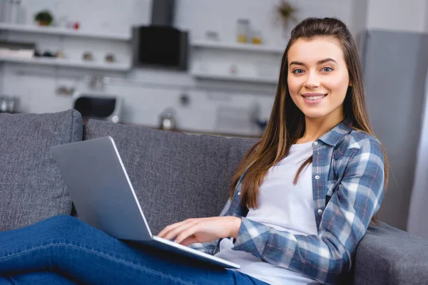 Atractiva chica feliz usando el ordenador portátil y sonriendo a la cámara - foto de stock