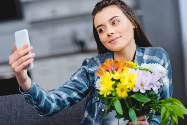 Hermosa joven sonriente sosteniendo flores y tomando selfie con teléfono inteligente en casa - foto de stock