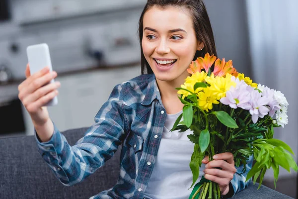 Ragazza felice che tiene bei fiori e prende selfie con smartphone a casa — Foto stock