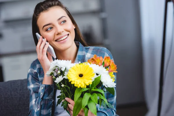 Улыбающаяся молодая женщина держит букет цветов и разговаривает со смартфоном — стоковое фото