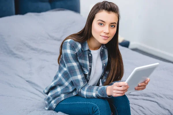 Усміхнена молода жінка сидить на ліжку і використовує цифровий планшет — стокове фото
