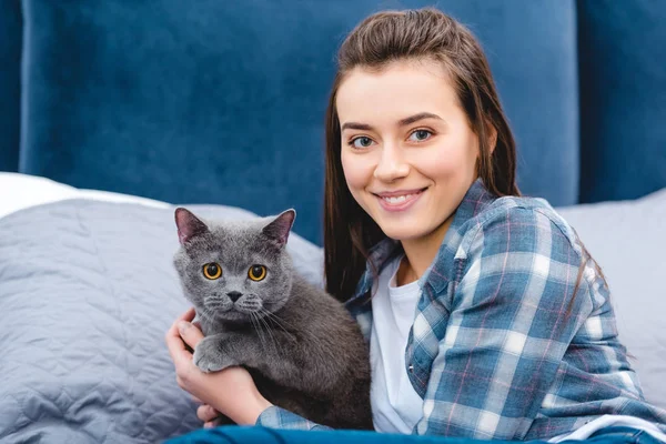 Hermosa feliz joven mujer abrazando gato y sonriendo a la cámara en el dormitorio - foto de stock