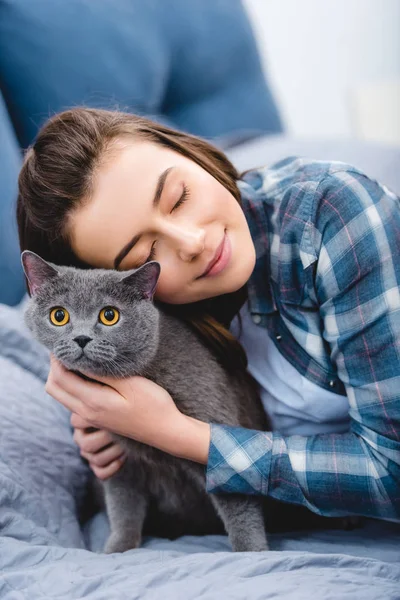 Happy girl with closed eyes hugging beautiful british shorthair cat on bed — Stock Photo