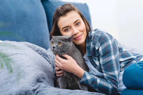 Menina feliz abraçando bonito gato cinza e sorrindo para a câmera no quarto — Fotografia de Stock