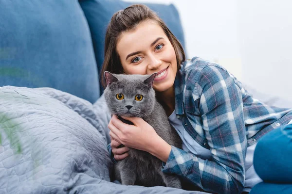 Feliz joven mujer abrazando lindo gris gato y sonriendo en cámara en dormitorio - foto de stock