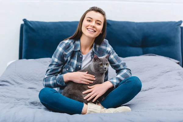 Chica feliz sentado en la cama con lindo británico taquigrafía gato y sonriendo a la cámara - foto de stock
