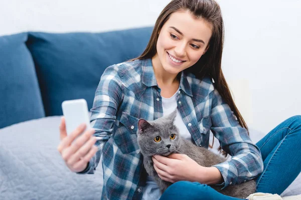 Heureuse jeune femme en utilisant smartphone et en prenant selfie avec chat gris mignon — Photo de stock