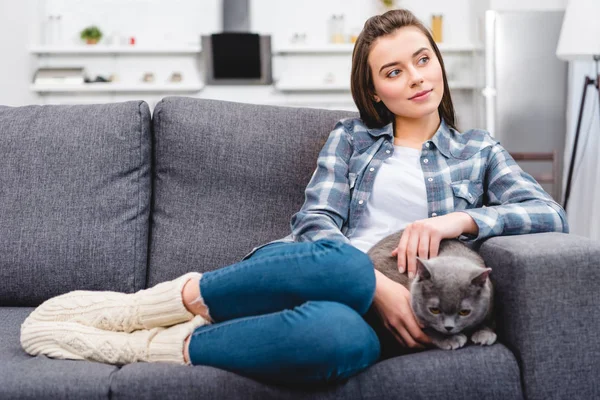 Hermosa chica sonriente sentado en el sofá con lindo gato gris - foto de stock