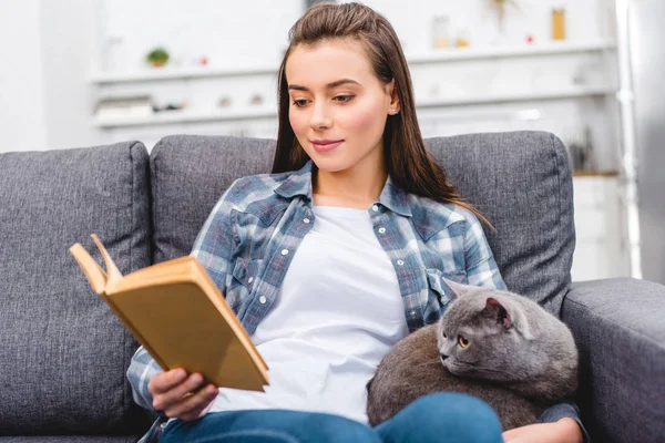 Souriant jeune femme lecture livre et assis avec chat sur canapé — Photo de stock