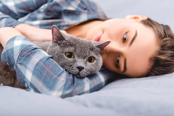 Красива щаслива дівчина лежить на ліжку з британським короткошерстим котом — стокове фото