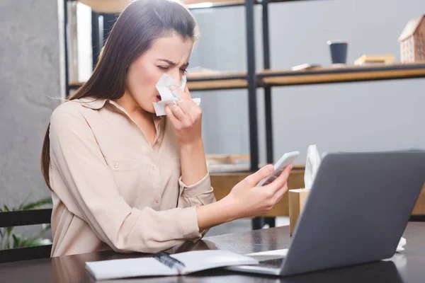 Young businesswoman holding smartphone and suffering from allergy at workplace — Stock Photo