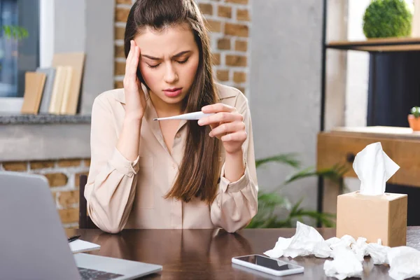 Sick young businesswoman holding thermometer and checking temperature in office — Stock Photo