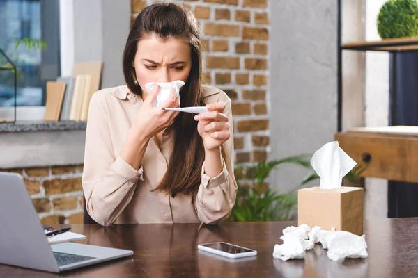 Sick young businesswoman blowing nose and holding thermometer in office — Stock Photo