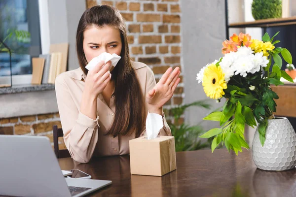 Jovem empresária assoprando nariz e olhando para flores enquanto sofre de alergia no local de trabalho — Fotografia de Stock