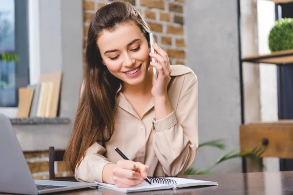 Smiling young businesswoman talking by smartphone at taking notes at workplace — Stock Photo