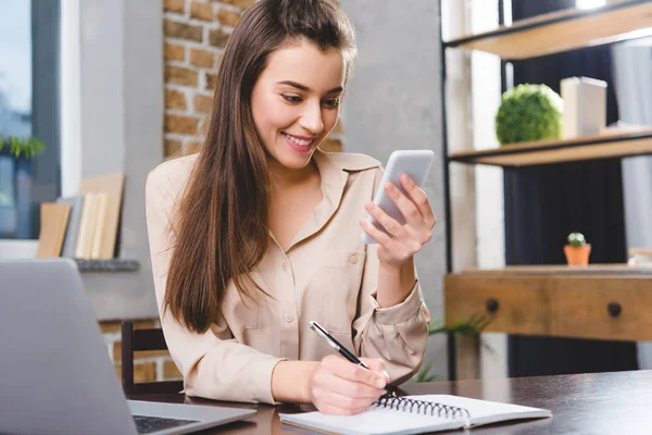 Beatiful smiling young businesswoman using smartphone while working in office — Stock Photo