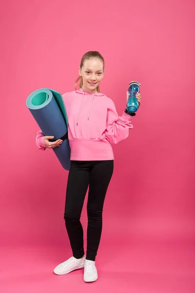 Adorable child holding fitness mat and sports bottle with water, isolated on pink — Stock Photo
