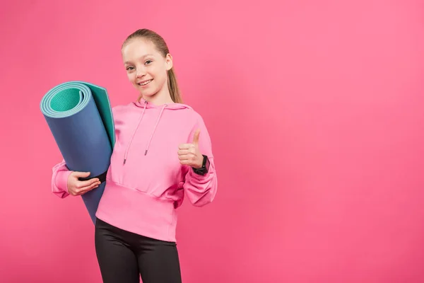 Femmina giovane che tiene il tappetino fitness e mostra pollice in alto, isolato su rosa — Foto stock