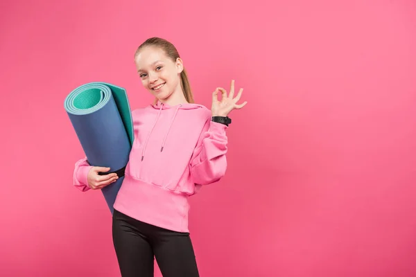 Счастливый ребенок, держащий фитнес-коврик и показывающий знак ОК, изолированный на розовом — стоковое фото
