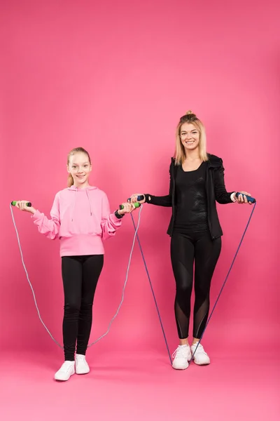 Madre e hija deportiva en ropa deportiva posando con cuerdas de saltar, aisladas en rosa - foto de stock