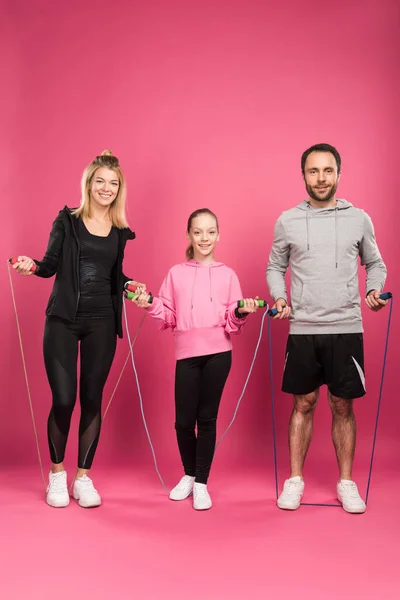 Parents and daughter in sportswear exercising with skipping ropes, isolated on pink — Stock Photo